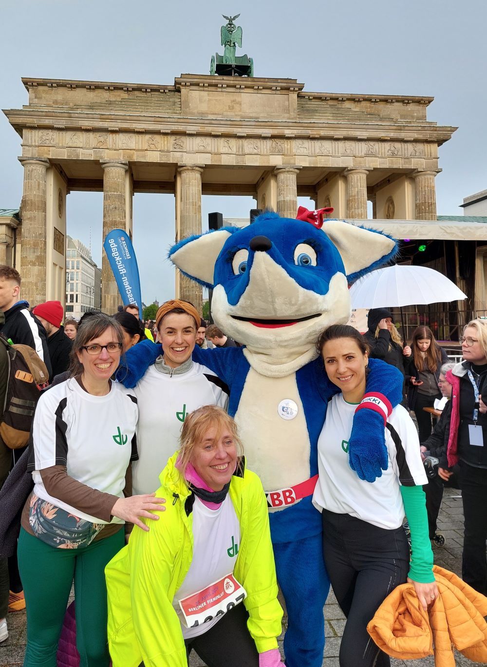 Das Bild ist ein Foto von 4 Kolleginnen die für den Deutschen Verein am Firmenlauf teilgenommen haben zusammen mit dem Maskottchen des Veranstalters. Das Maskottchen ist ein blauweißer Fuchs. Im Hintergrund sind weitere Menschen und das Brandenburger Tor zu sehen.
