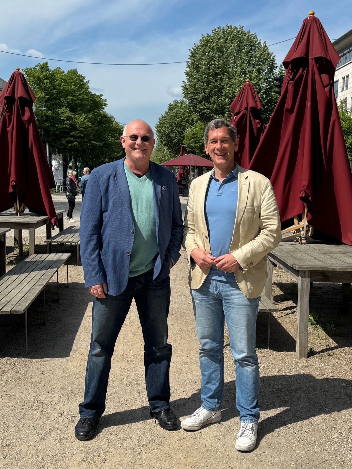 Foto: Auf dem Foto sind Michael Löher und Jens Kamieth zu sehen, die nebeneinander auf einer Terrasse stehen. Im Hintergrund stehen weinrote Sonnenschirme, Holztische und Holzstühle.