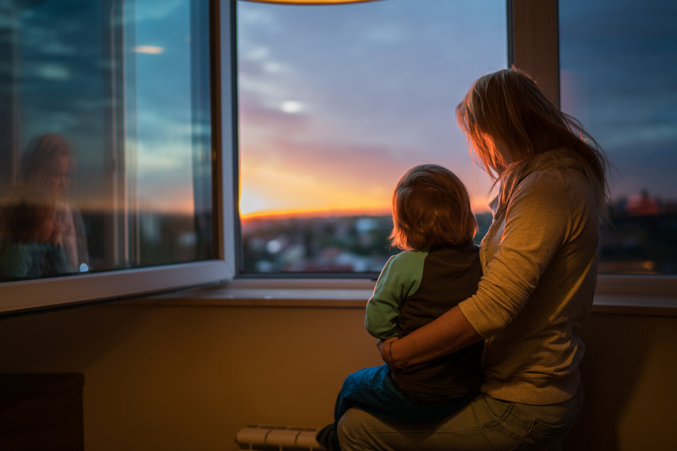 Foto einer Frau, sitzend vor einem Fenster mit ihrem Kind auf dem Arm. –