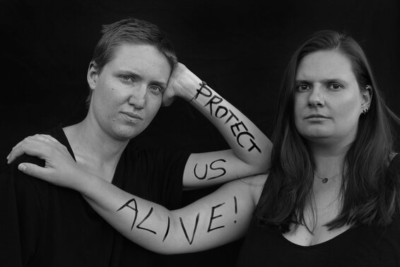 Bild in schwarz-weiß von zwei Frauen mit der Aufschrift "protect us alive!" auf den Armen der beiden Frauen. – 