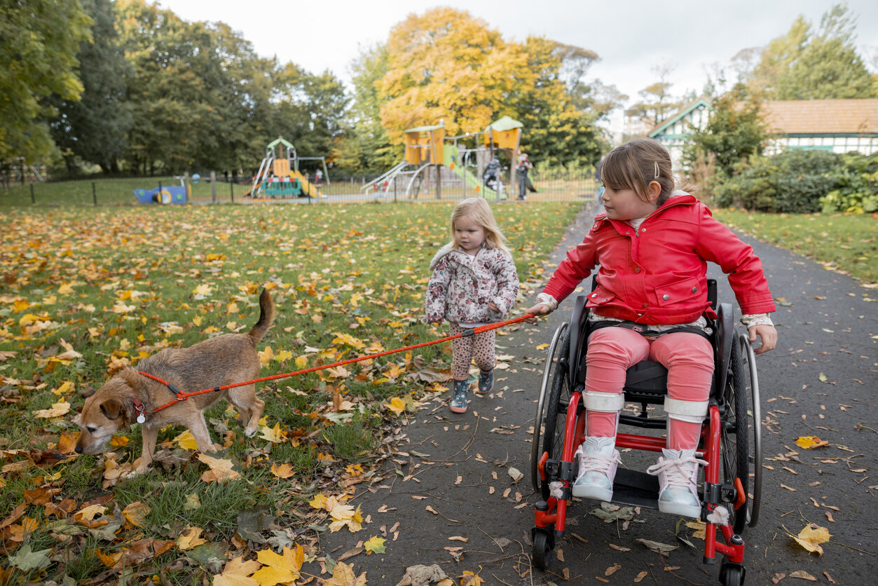 Eine Vorderansicht von zwei Schwestern, die mit ihrem Hund in einem öffentlichen Park in Newcastle upon Tyne im Nordosten Englands spazieren gehen, sie sind mit ihren Eltern unterwegs und genießen es, an einem klaren Herbsttag im Park zu spielen. –