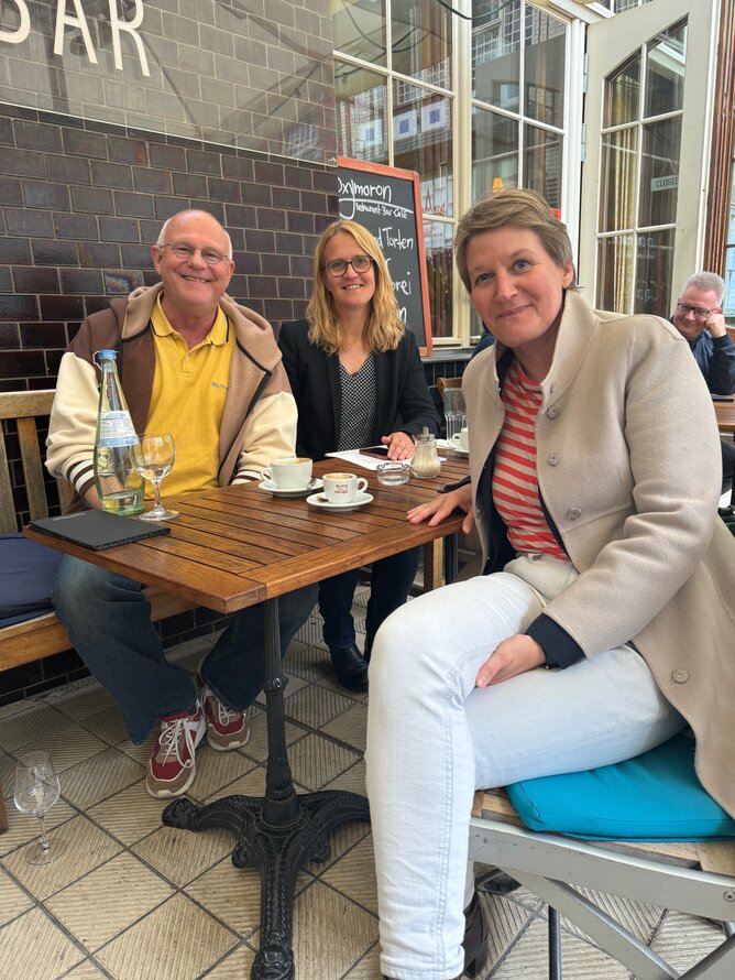Auf dem Foto sitzen Michael Löher, Nora Schmidt und Evelin Schulz an einem Tisch in einem Café. Alle drei schauen in die Kamera. –