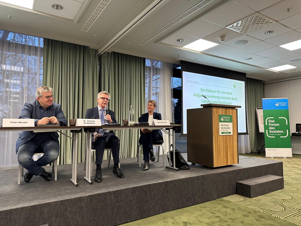Auf dem Foto sitzen von links Peter Renzel, Dr. Thomas de Maizière und DV-Geschäftsführerin Nora Schmidt auf einer Bühne. Dr. de Maizière hält dabei ein Mikrofon in der Hand und spricht in Richtung der Zuhörer*innen. – 
