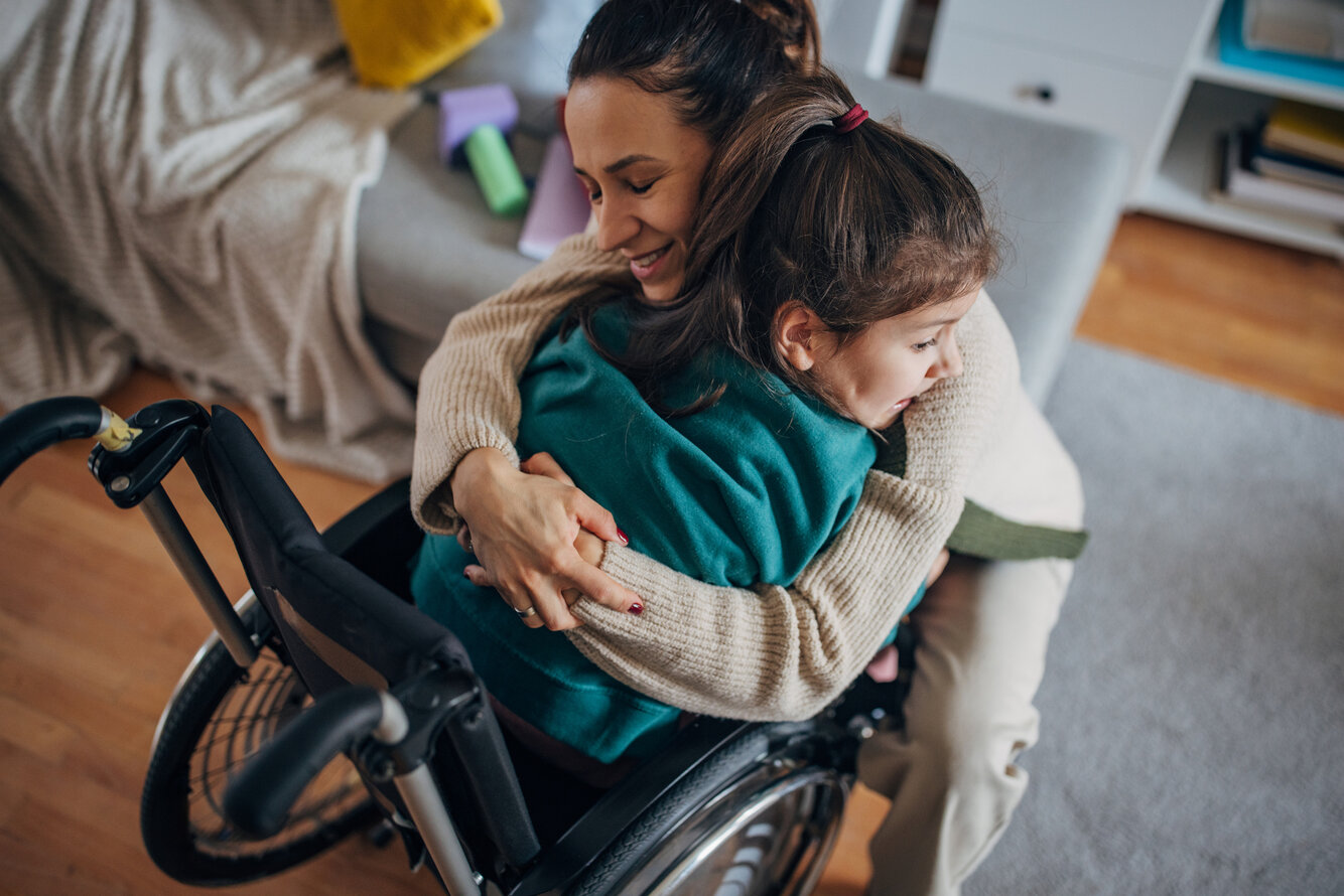 Foto eines Kindes im Rollstuhl, das von seiner Mutter umarmt wird. – 
