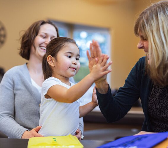 Auf dem Foto sind 2 Frauen und ein Kind zu sehen. Die Personen befinden sich in einem Wohnzimmer, das Kind befindet sich in der Mitte des Bildes. Die beiden Frauen sind neben dem Kind zu sehen und lächeln. – 