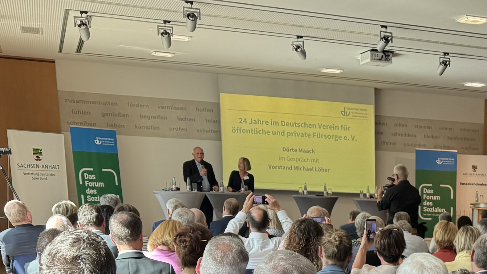 Das Bild zeigt Michael Löher und die Moderatorin auf der Bühne stehen. Im Hintergrund ist die Leinwand. Links und rechts der Bühne steht ein Roll-up. Vorne im Bild sind die Teilnehmenden zu sehen mit dem Rücken der Aufnahme zugewandt. –
