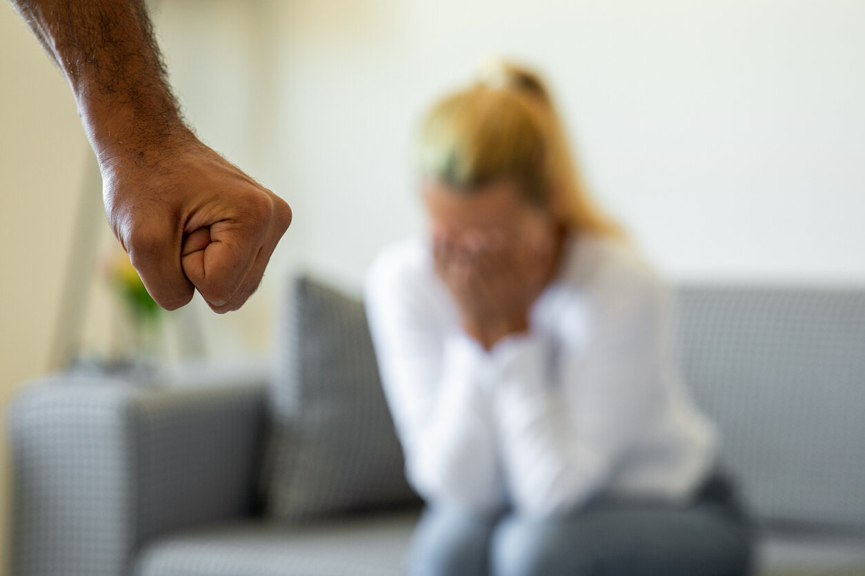 Eine Frau auf dem Sofa mit den Händen vors Gesicht, im Vordergrund eine geballte Faust. –