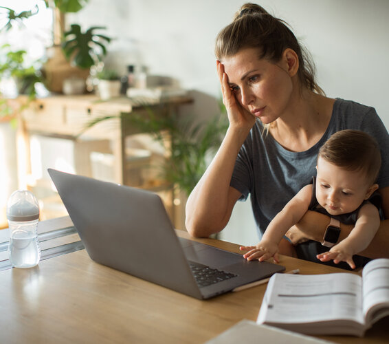 Auf dem Bild zu sehen ist eine Frau, die einen Säugling auf dem Arm hat, während sie am Schreibtisch vor einem Laptop sitzt. – 