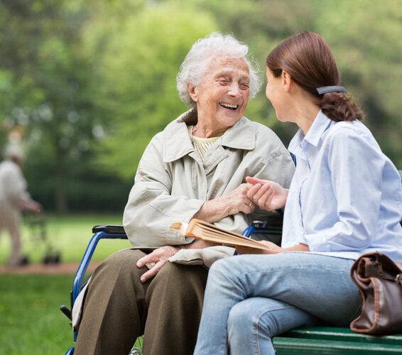 Im Vordergrund des Fotos sind eine junge und eine alte Frau zu sehen, die auf einer Parkbank sitzen. Die Blicke der beiden Personen richten sich einander zu und die ältere Frau lächelt und strahlt Freude aus. Im Hintergrund ist ein Park zu sehen und eine Person mit Rollator. – 