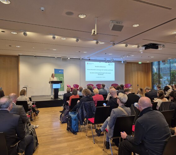 Foto eines Vortrags beim Forum Monetäre Leistungen. Im Vordergrund sind die Zuschauer*innen zu sehen, deren Blick nach vorne, weg von der Kamera zur Bühne hin gerichtet ist. Auf der Bühne steht eine Rednerin an einem Pult und präsentiert ihren Vortrag. – 
