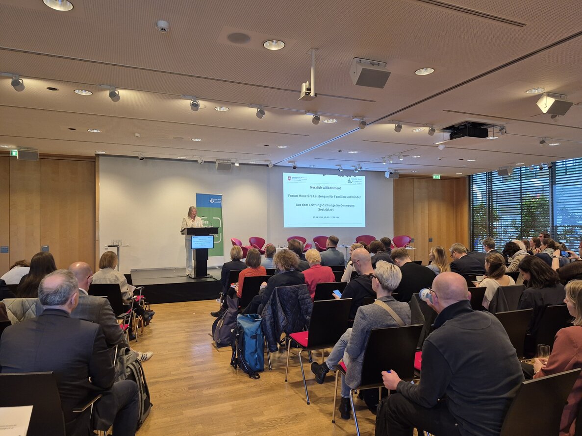 Foto eines Vortrags beim Forum Monetäre Leistungen. Im Vordergrund sind die Zuschauer*innen zu sehen, deren Blick nach vorne, weg von der Kamera zur Bühne hin gerichtet ist. Auf der Bühne steht eine Rednerin an einem Pult und präsentiert ihren Vortrag. – 