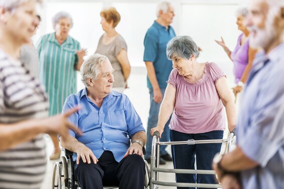 Das Bild zeigt eine Situation in einem Pflegeheim. In der Mitte unterhalten sich ein Mann, der in einem Rollstuhl sitzt und eine Frau mit Gehhilfe. An den Rändern des Bildes sind mehrere Senior*innen zu sehen. – 